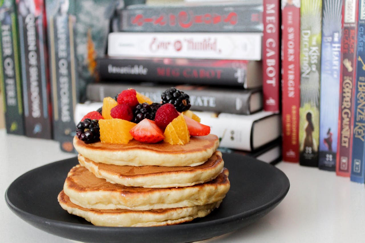 who-we-are Stack of pancakes topped with fresh berries and oranges against a backdrop of books.