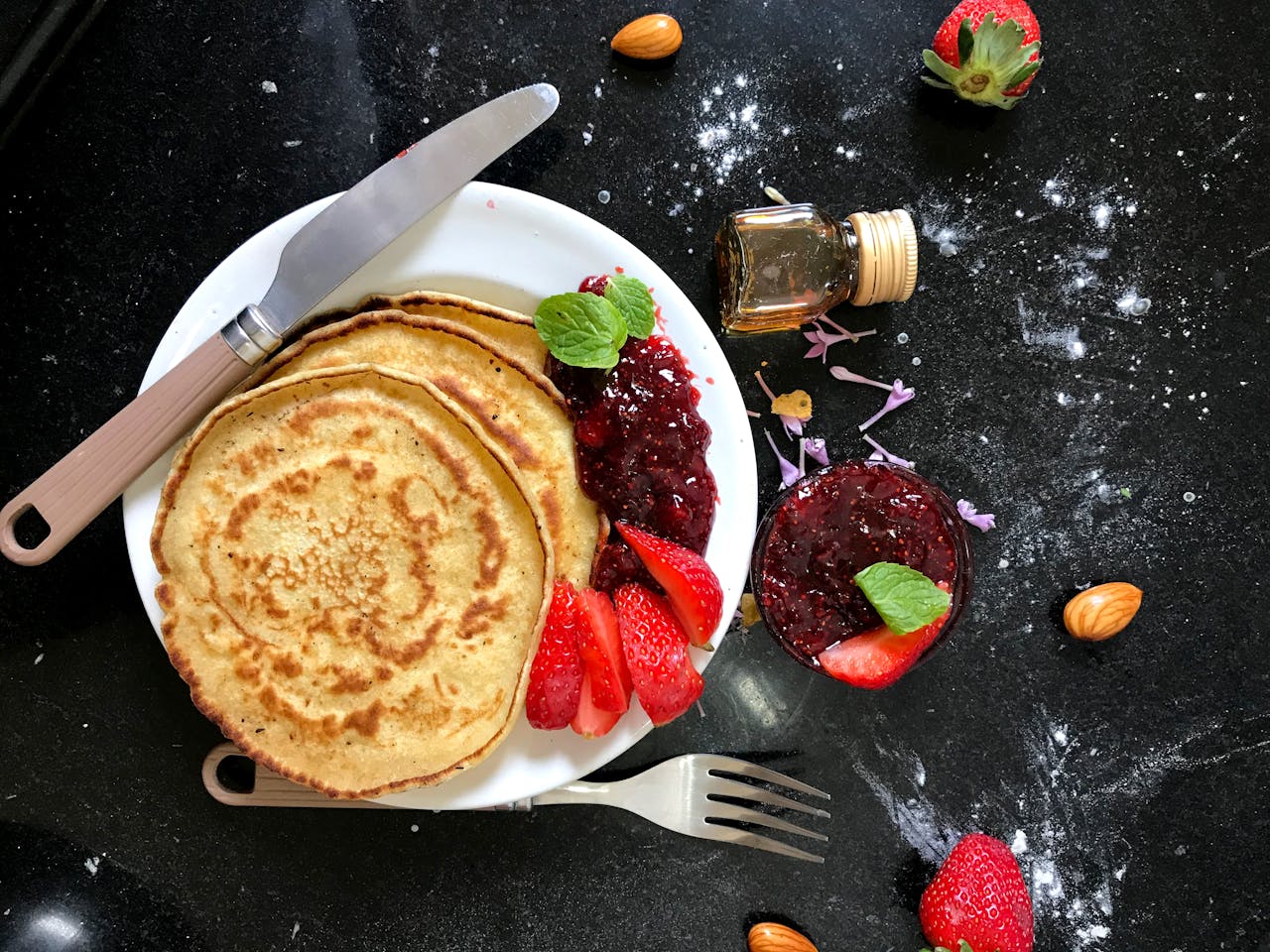why-choose-us Top view of pancakes with strawberry jam, fresh strawberries, almonds, and honey on a dark kitchen counter.