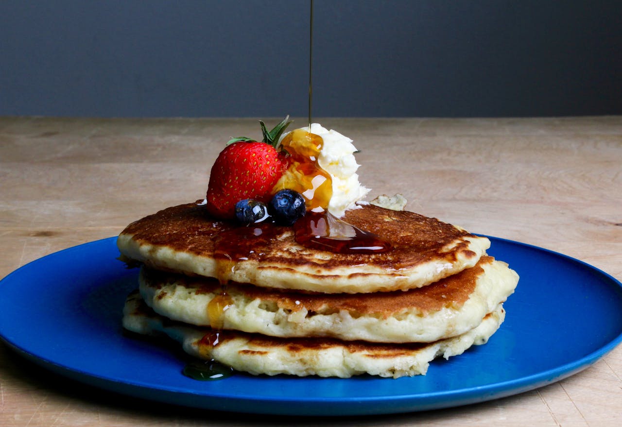 services-01 Stack of fluffy pancakes topped with syrup, strawberries, blueberries, and cream on a blue plate.
